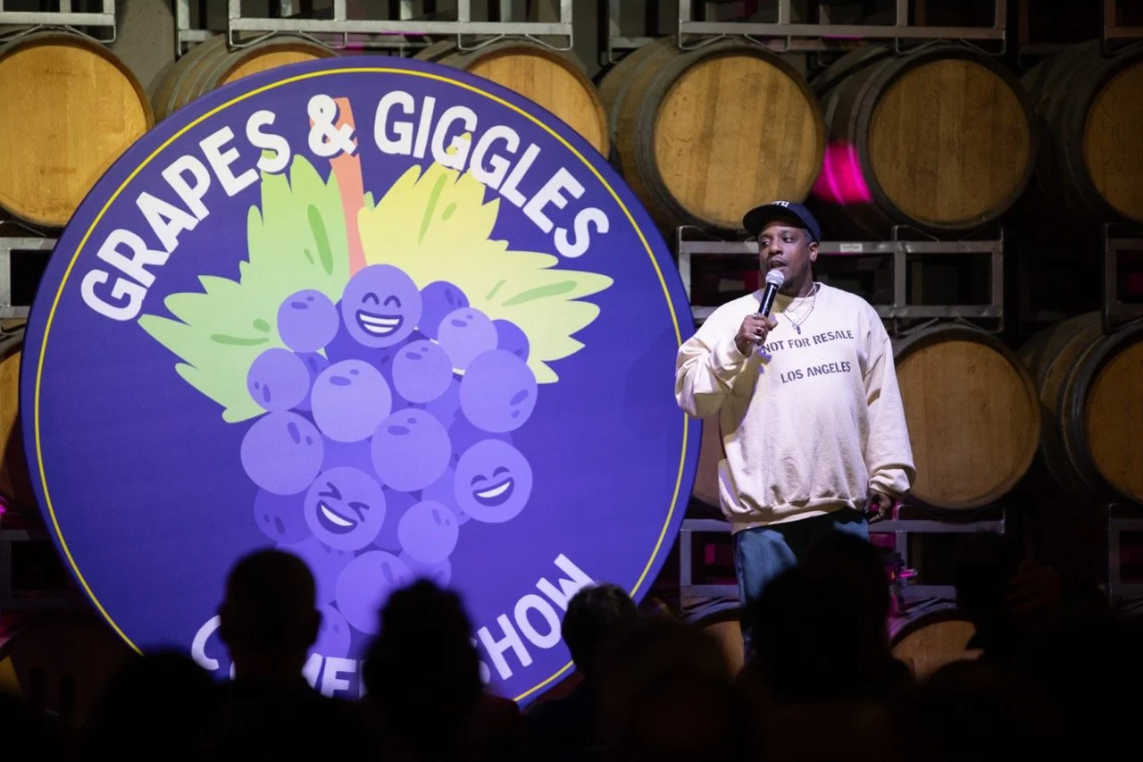 Comedian performing stand-up comedy in front of a "Grapes & Giggles" sign with smiling grape illustrations, with audience in foreground and wine barrels in the background.