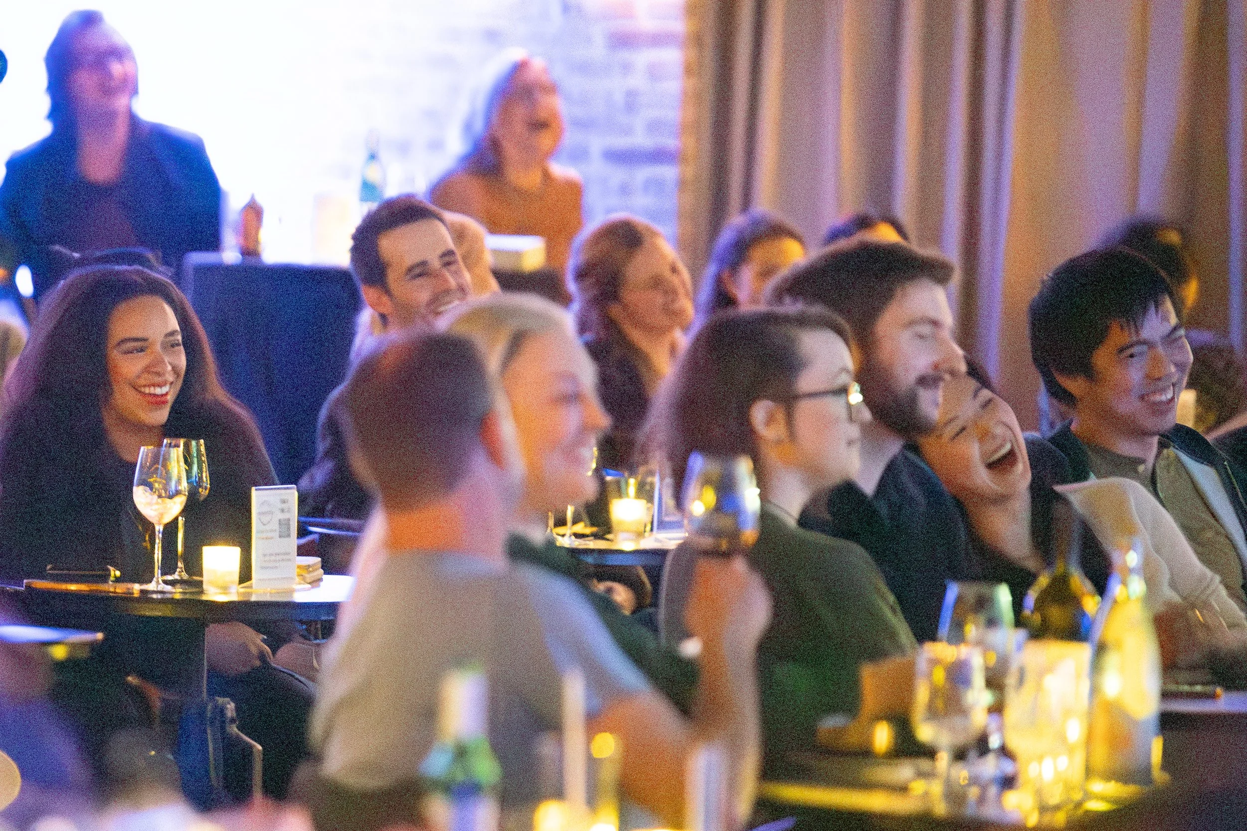A group of people seated at a comedy show, laughing and enjoying themselves in a dimly lit room with tables and wine glasses.