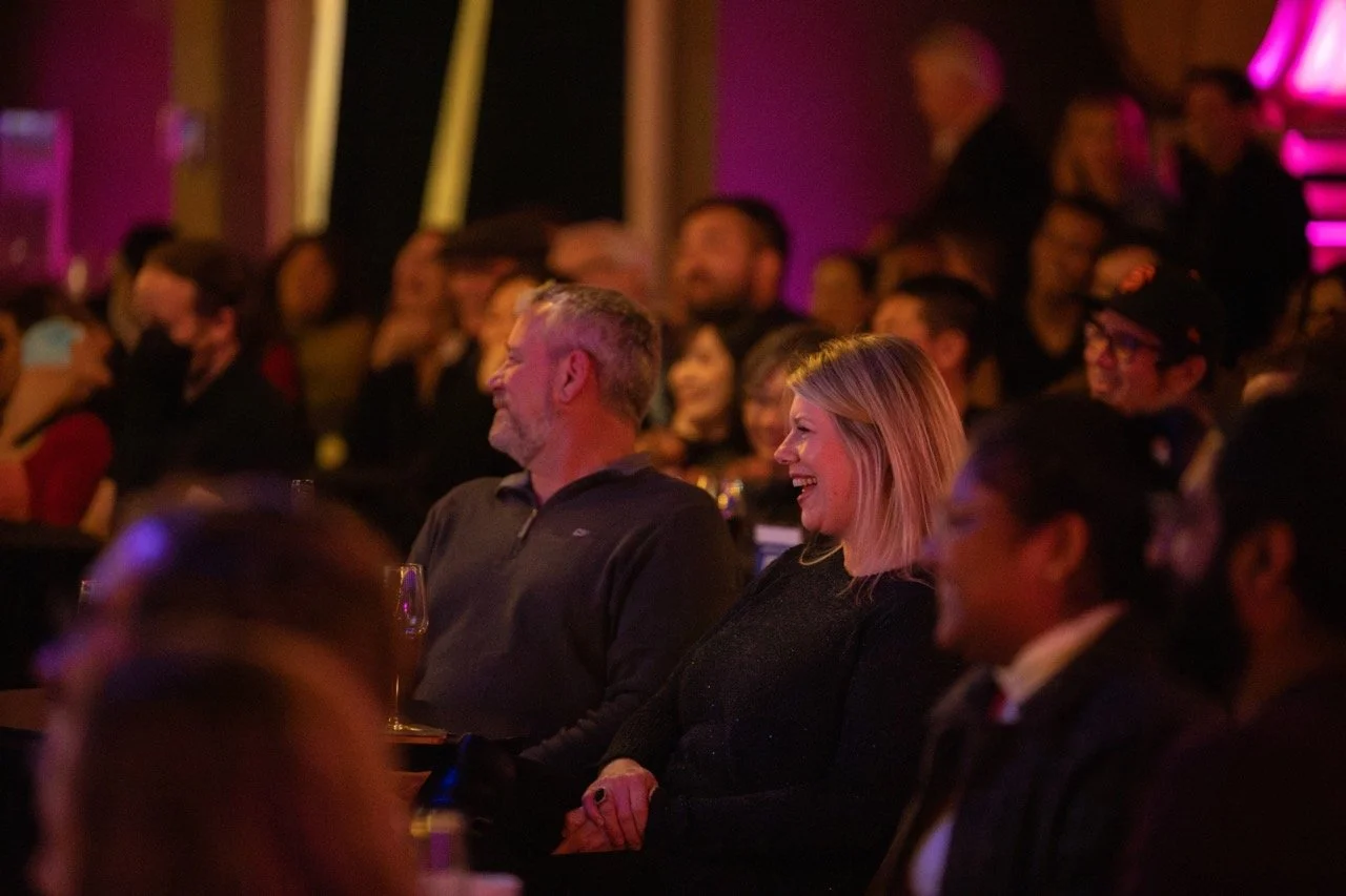 People seated in an audience, smiling and enjoying a performance, with a dimly lit, warm and festive ambiance.