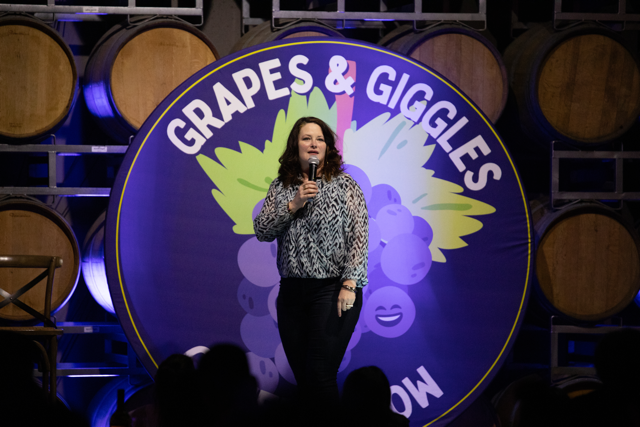 Comedian performing at the Grapes & Giggles comedy show with a backdrop of wine barrels.
