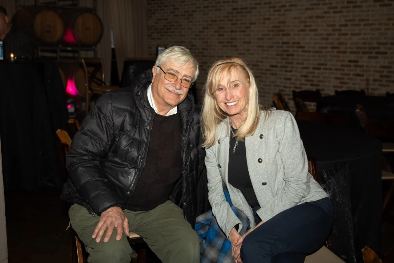 An older man and woman smiling, seated indoors next to each other, with a brick wall and barrels in the background. The man is wearing glasses and a black jacket, and the woman has long blonde hair and a light-colored coat.