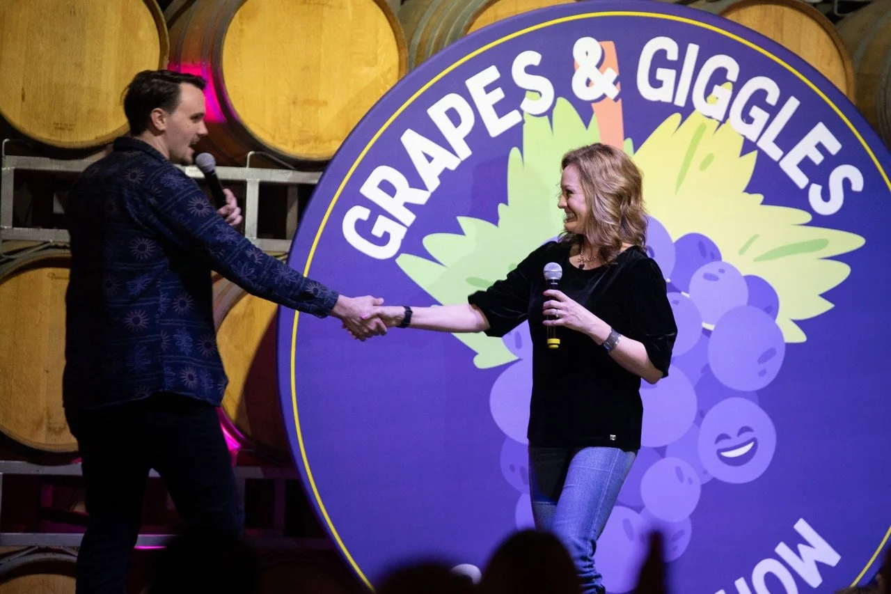 Two people on stage at a comedy and wine event called "Grapes & Giggles," holding microphones and shaking hands. Background shows wine barrels and event signage.
