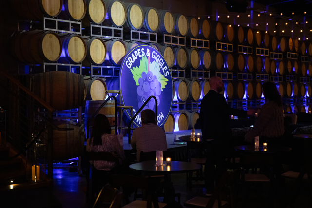 Barrel room with stage and sign for comedy event "Grapes & Giggles," tables with candles