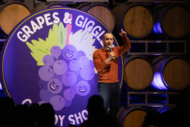 Comedian performing at 'Grapes & Giggles Comedy Show' with barrels in the background.