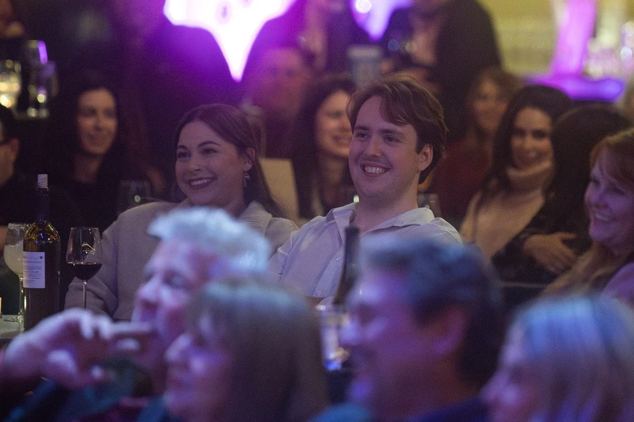 Smiling people sitting at a comedy show or event with drinks on the table.