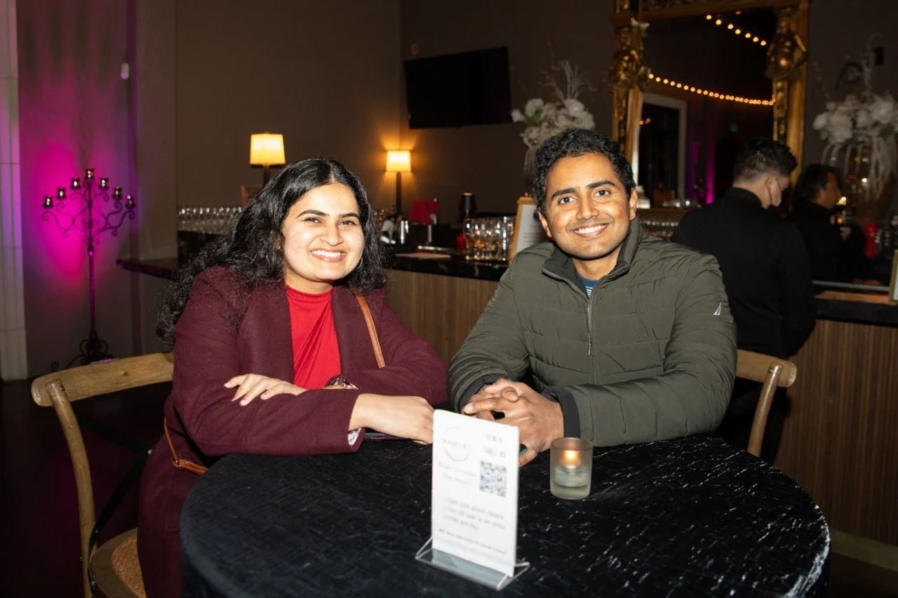 Two people smiling at a candle-lit table in a dimly lit room with a bar in the background, featuring decorative elements and warm lighting.
