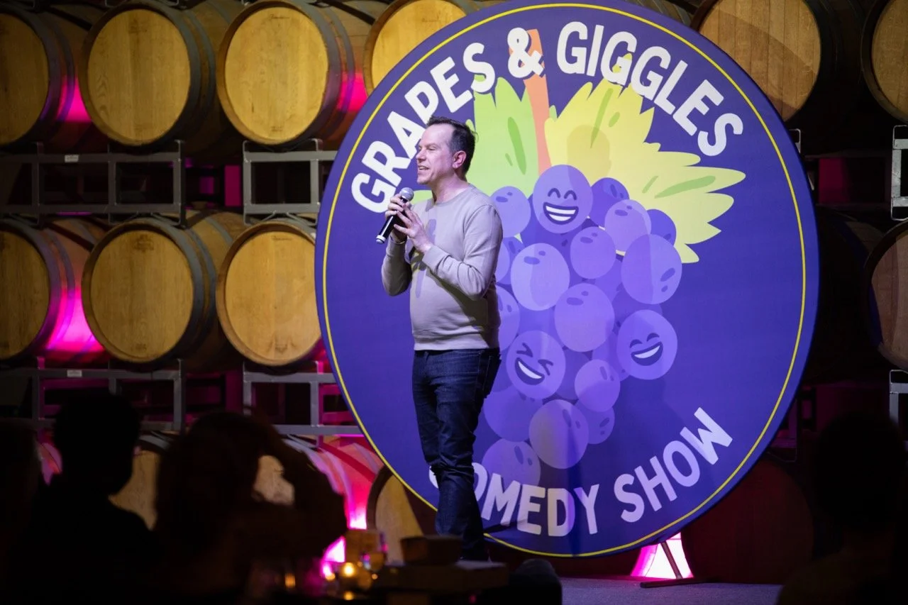 Comedian performing at "Grapes & Giggles Comedy Show" with wine barrels in background.