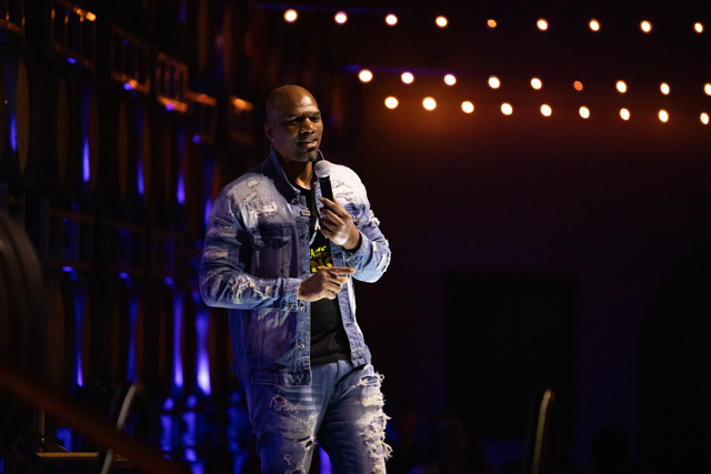 Comedian performing on stage, holding a microphone, wearing denim jacket and jeans, with string lights and audience in background.