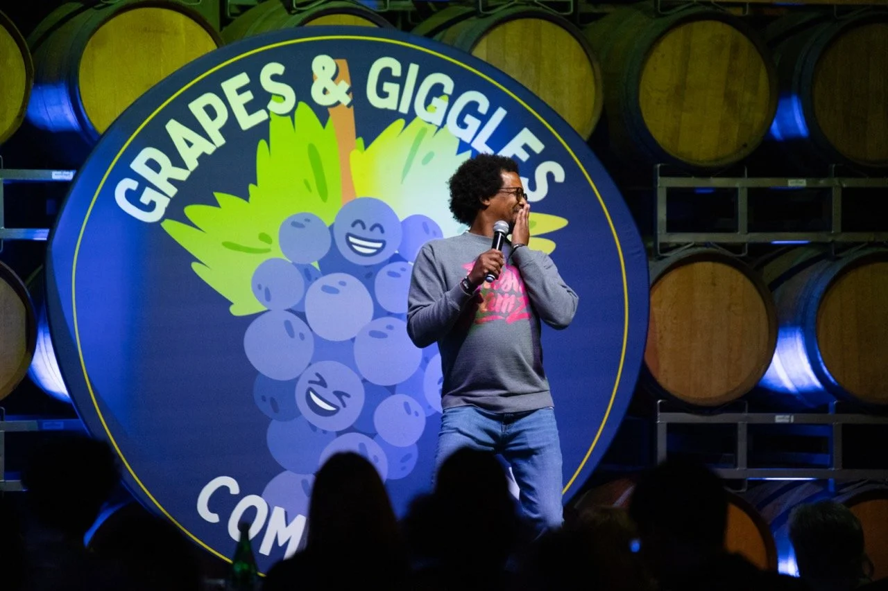 Comedian performing on stage at "Grapes & Giggles" event with wine barrels in the background.