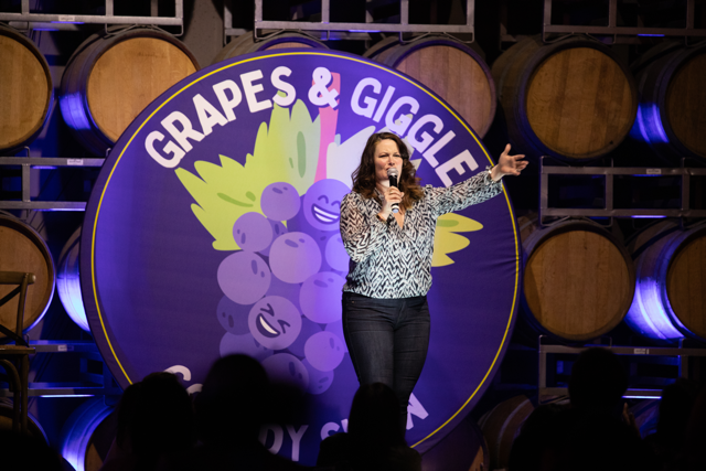 Comedian performing on stage with a "Grapes & Giggle Comedy Show" backdrop, surrounded by wine barrels.