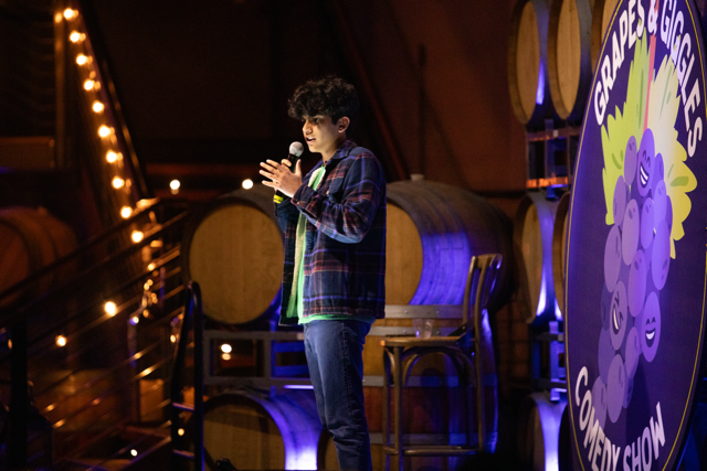 Comedian performing on stage with microphone at "Grapes and Giggles Comedy Show," surrounded by wine barrels and soft lighting.