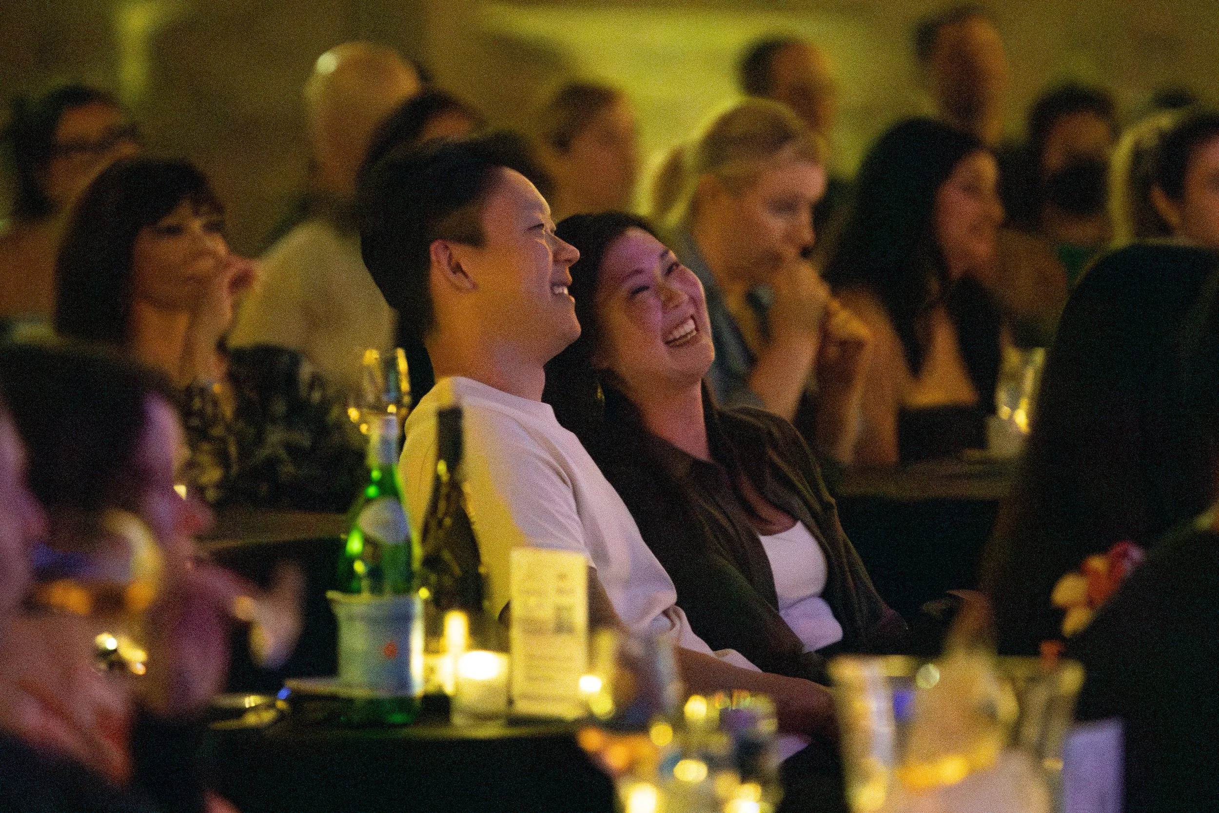 People laughing and enjoying a live event in a dimly lit venue, with bottles and glasses on the table.