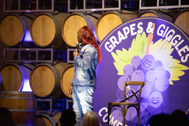 A comedian performs on stage with a microphone, wearing a denim outfit with red hair. The stage backdrop reads "Grapes & Giggles Comedy" with a grape-themed design. Wooden barrels are stacked in the background, suggesting a winery or barrel room sett