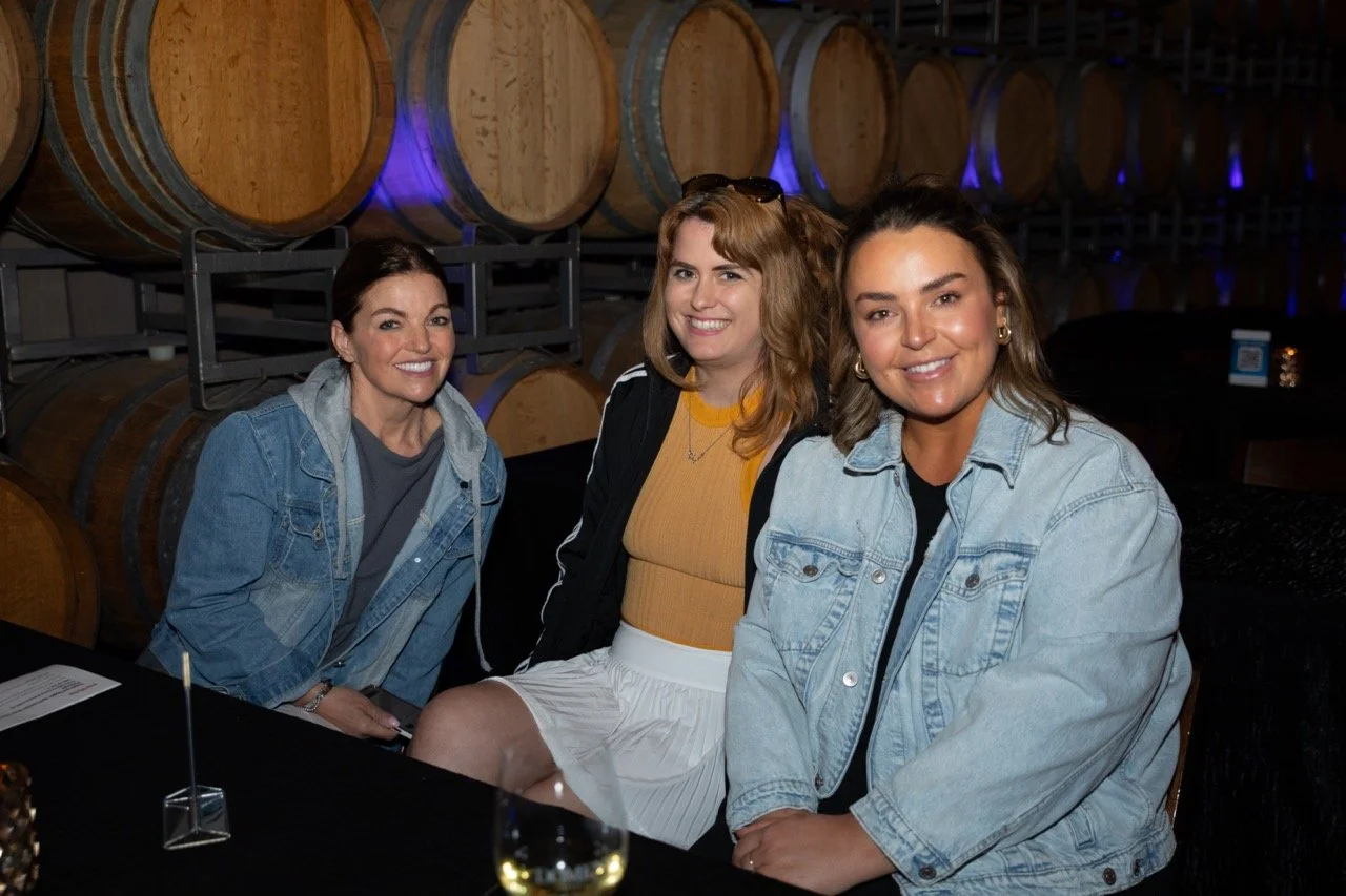 Three women sitting together smiling in front of wine barrels in a winery setting.