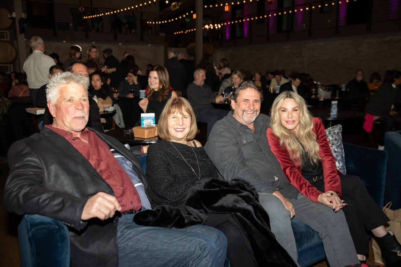 Group of four people sitting at a social event, with tables and others in the background.