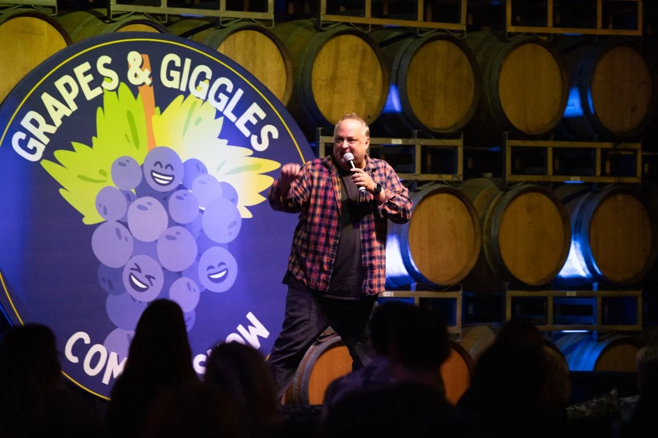 Comedian performing at Grapes & Giggles comedy show, wooden barrels in background.