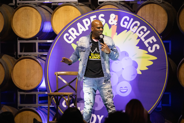 Comedian performing on stage labeled 'Grapes & Giggles' with wine barrels in the background.