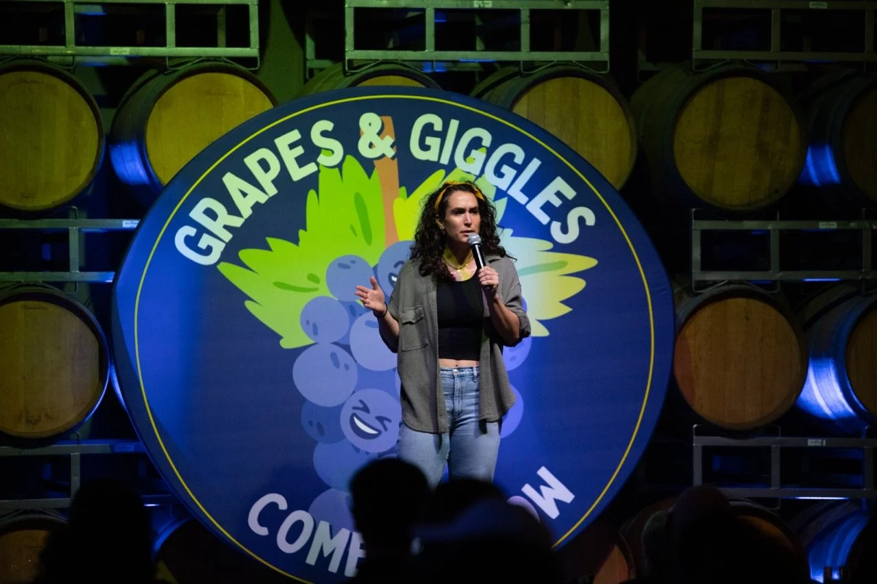 Comedian performing on stage at "Grapes & Giggles Comedy Show"