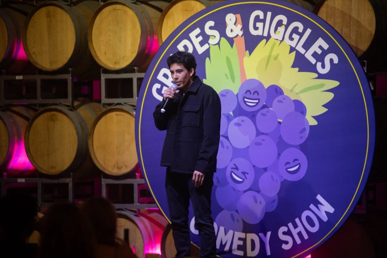 A comedian performing on stage at the "Grapes & Giggles" comedy show, standing in front of a backdrop with a cartoon grape design. Wine barrels are visible in the background with pink lighting.