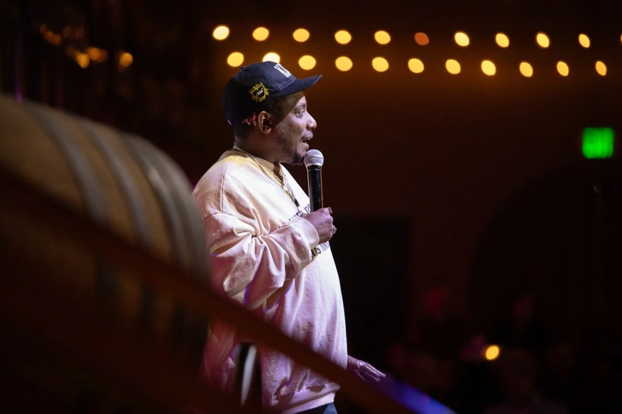 Comedian performing on stage holding a microphone, wearing a cap and light-colored sweater. Audience and string lights visible in the background.