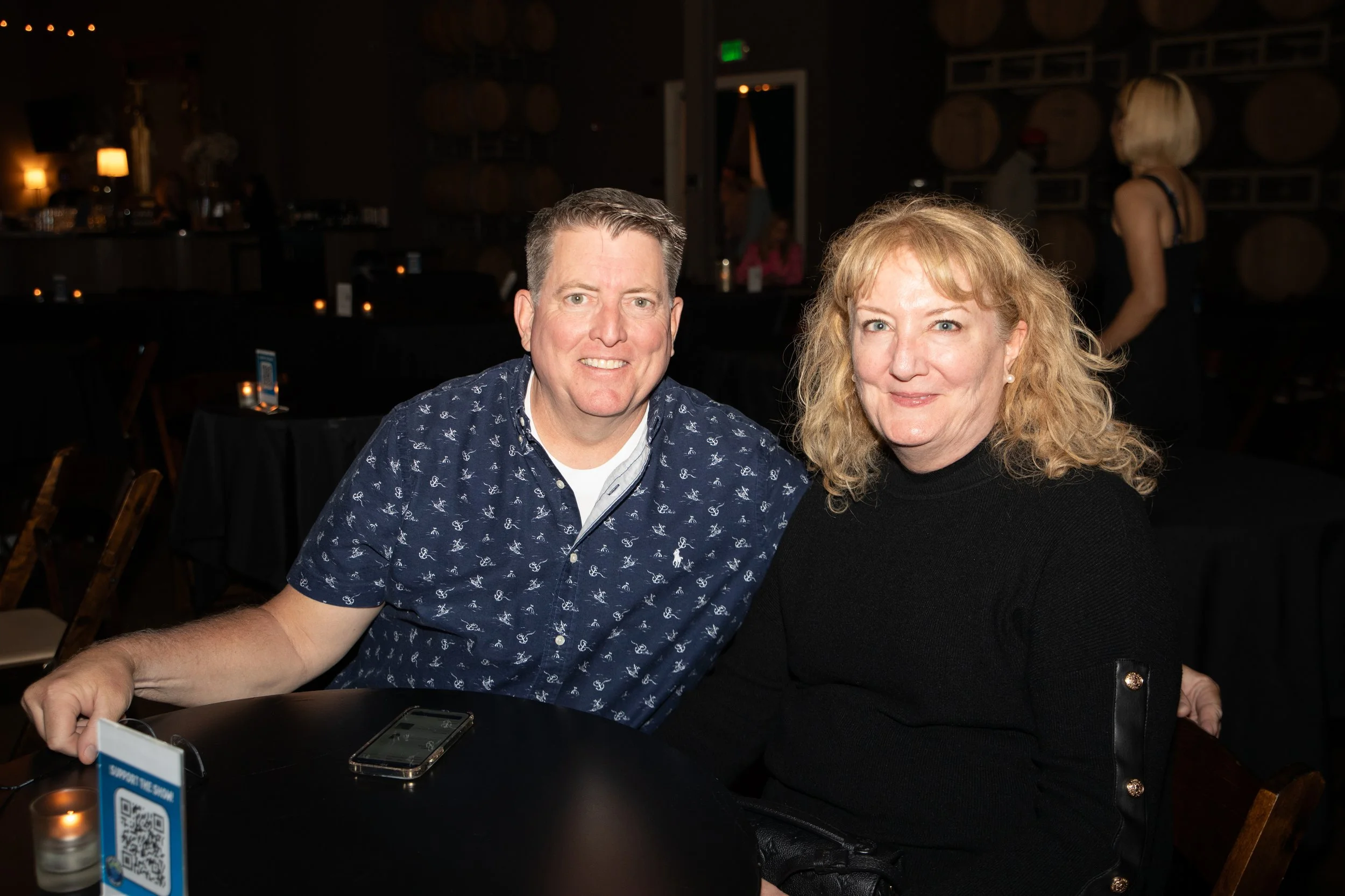 A couple seated at a table in a dimly lit venue, smiling at the camera, with a QR code display and candle on the table.