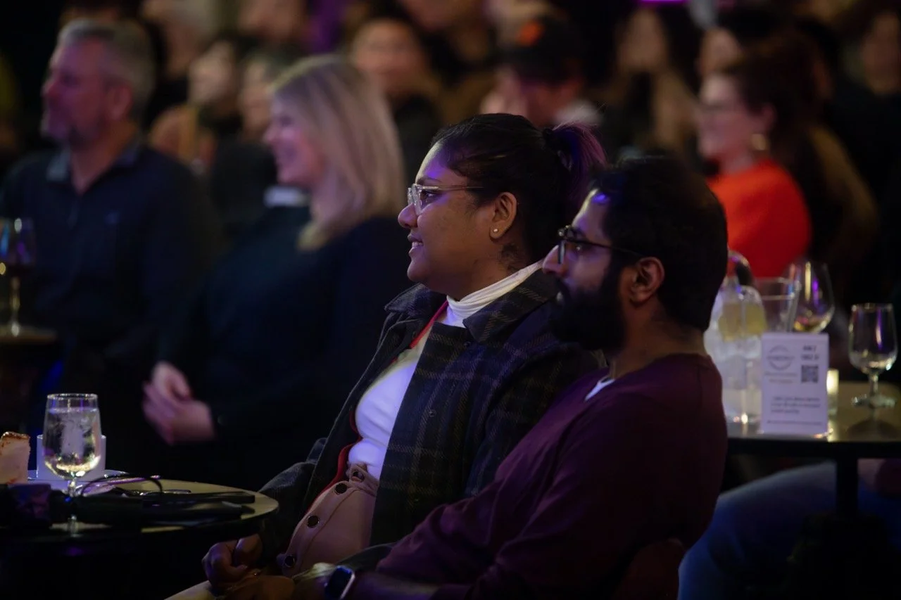 Audience members in a dimly lit event setting, seated at tables with drinks, attentively watching something off-camera.