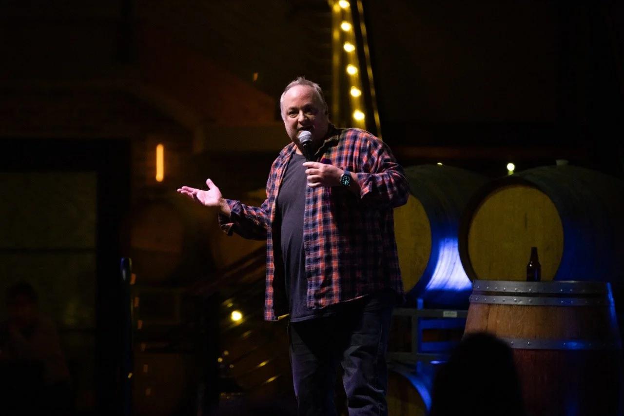 Comedian performing on stage holding a microphone, with wine barrels in the background.