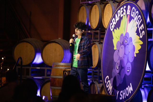 Comedian performing stand-up at the Grapes & Giggles Comedy Show, with wine barrels in the background and a colorful event sign.