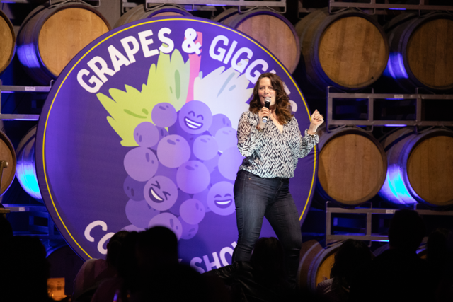 A comedian performing on stage at the "Grapes & Giggles" comedy show, standing in front of a backdrop featuring cartoon grapes. The venue has wine barrels stacked in the background.