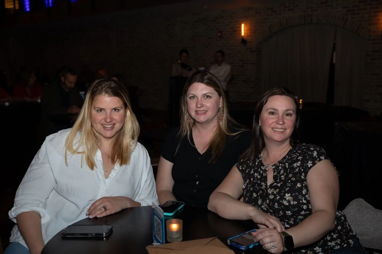 Three women sitting at a table in a dimly lit room, smiling at the camera, with drinks and phones on the table.