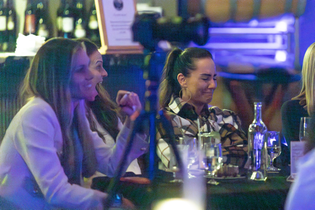 A group of women seated at a table, laughing at an event, with wine bottles in the background.