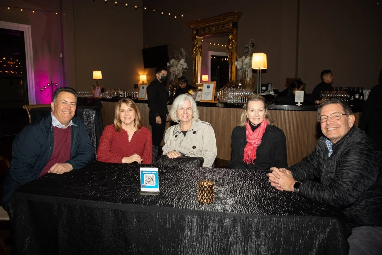 Group of five people sitting at a table in a dimly lit bar or lounge, with a decorated bar and attendants in the background.