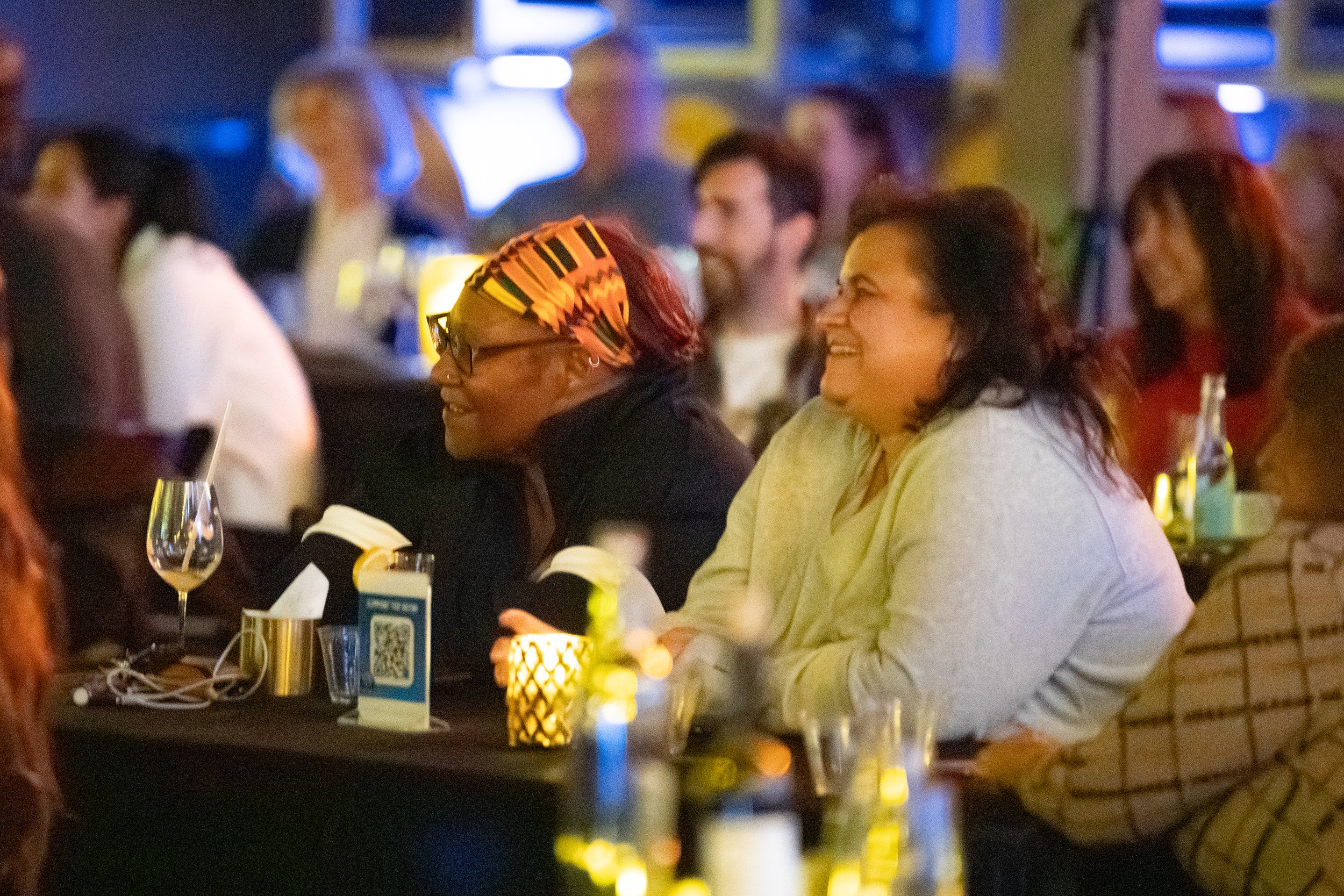 Audience members enjoying a comedy show, laughing and smiling, with drinks on the table.