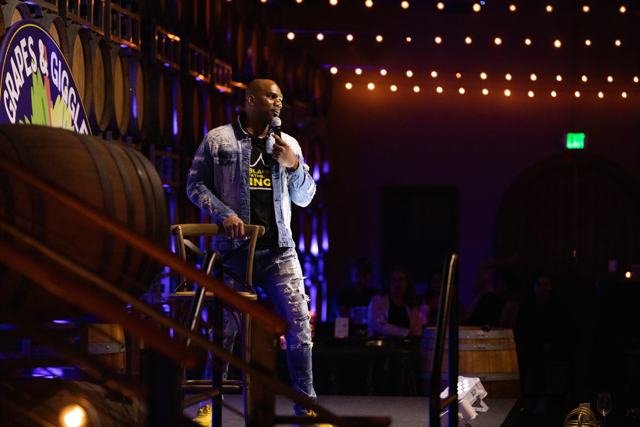 Comedian performing on stage in a dimly lit venue with barrels and string lights in the background.