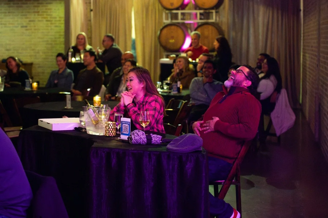 People laughing and enjoying themselves at a comedy show in a cozy venue with candlelit tables and barrel decorations.