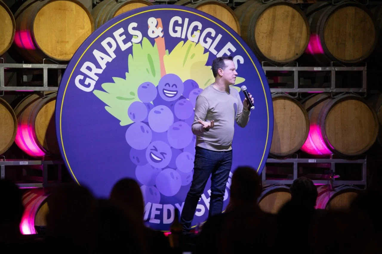 Comedian performing at Grapes & Giggles comedy show with wine barrels in the background.