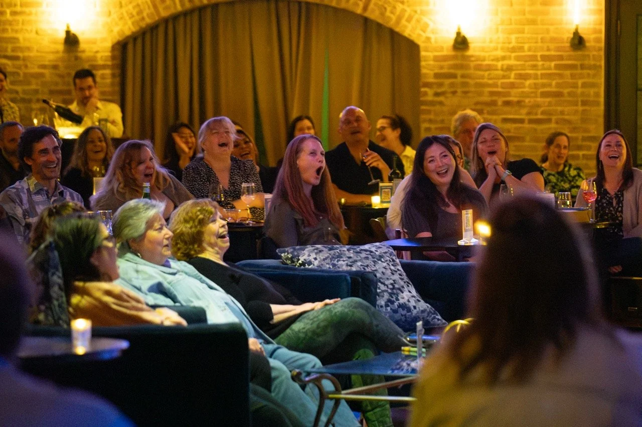 Audience laughing at a comedy show, seated in a dimly lit venue with brick walls and candles on tables.