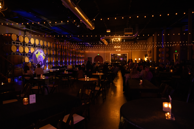 Dimly lit event space with tables and chairs, featuring wine barrels lining the walls, string lights overhead, and candles on the tables. A stage with a sign and a gathering of people is visible.