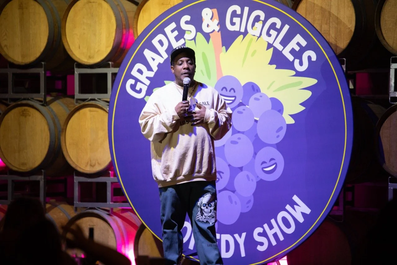 Comedian performing on stage in front of "Grapes & Giggles Comedy Show" sign, with barrels in the background.