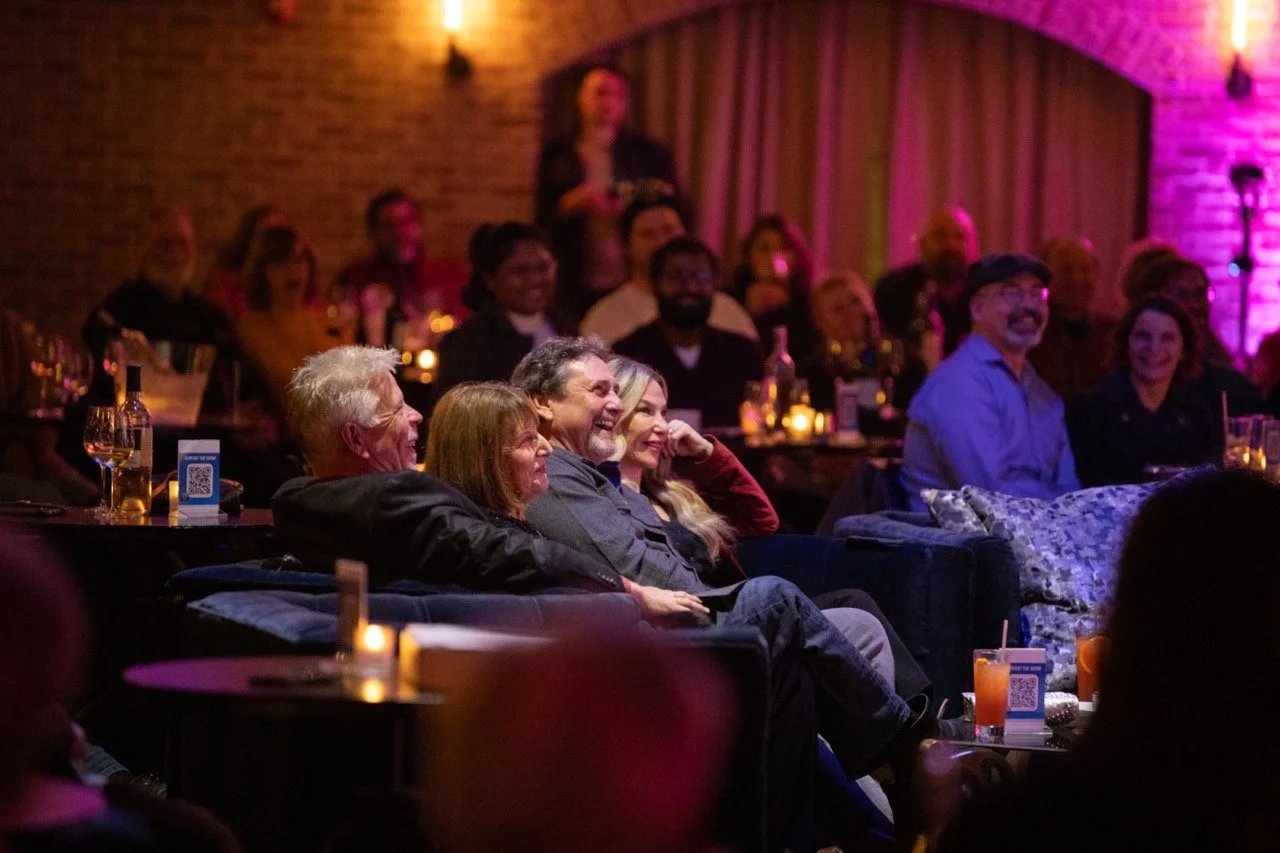 Audience enjoying a performance in a dimly lit comedy club, seated on couches with drinks and lit candles on tables.
