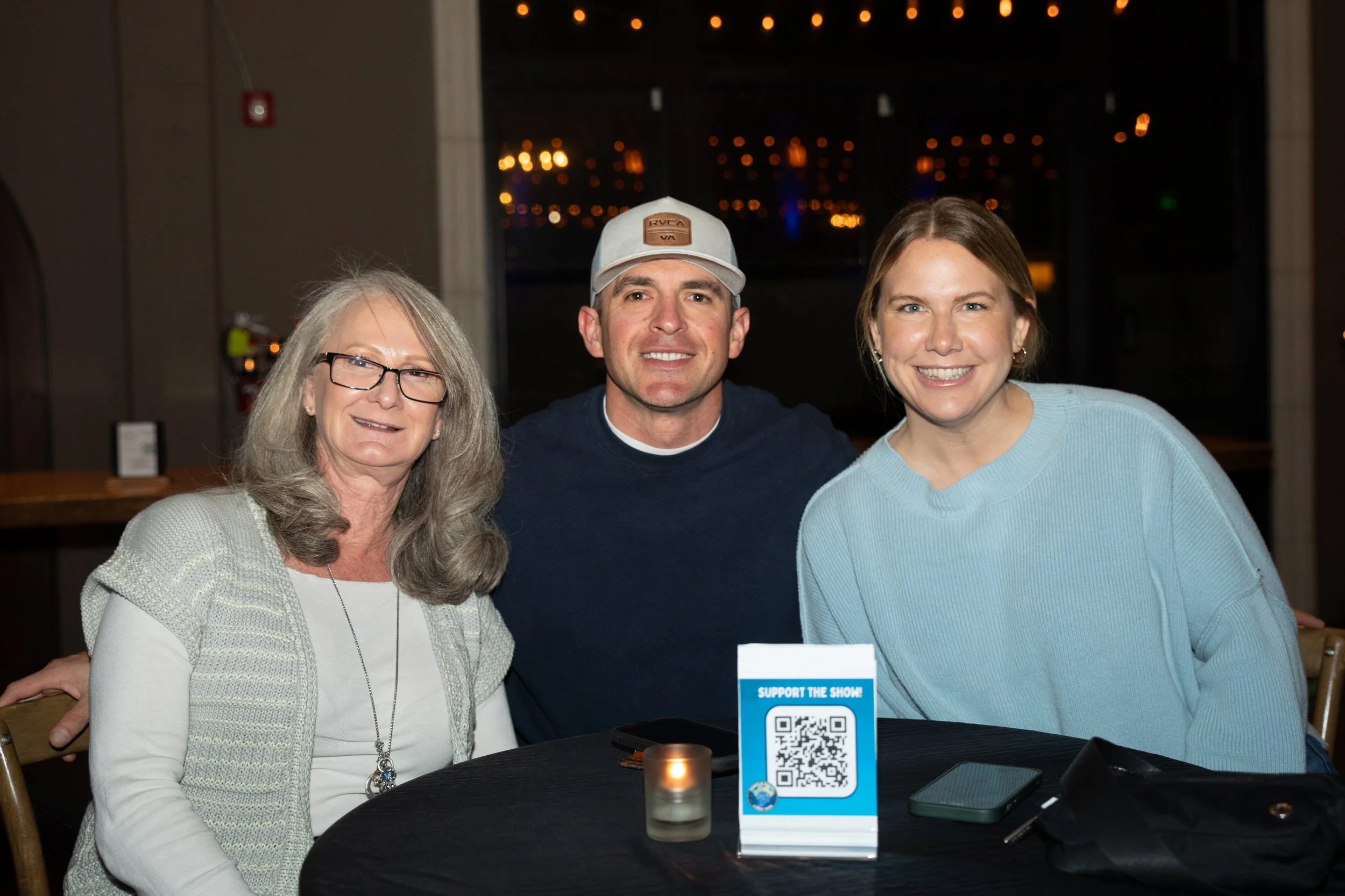 Three people sitting at a table, smiling, with a "Support the Show" sign in front of them.