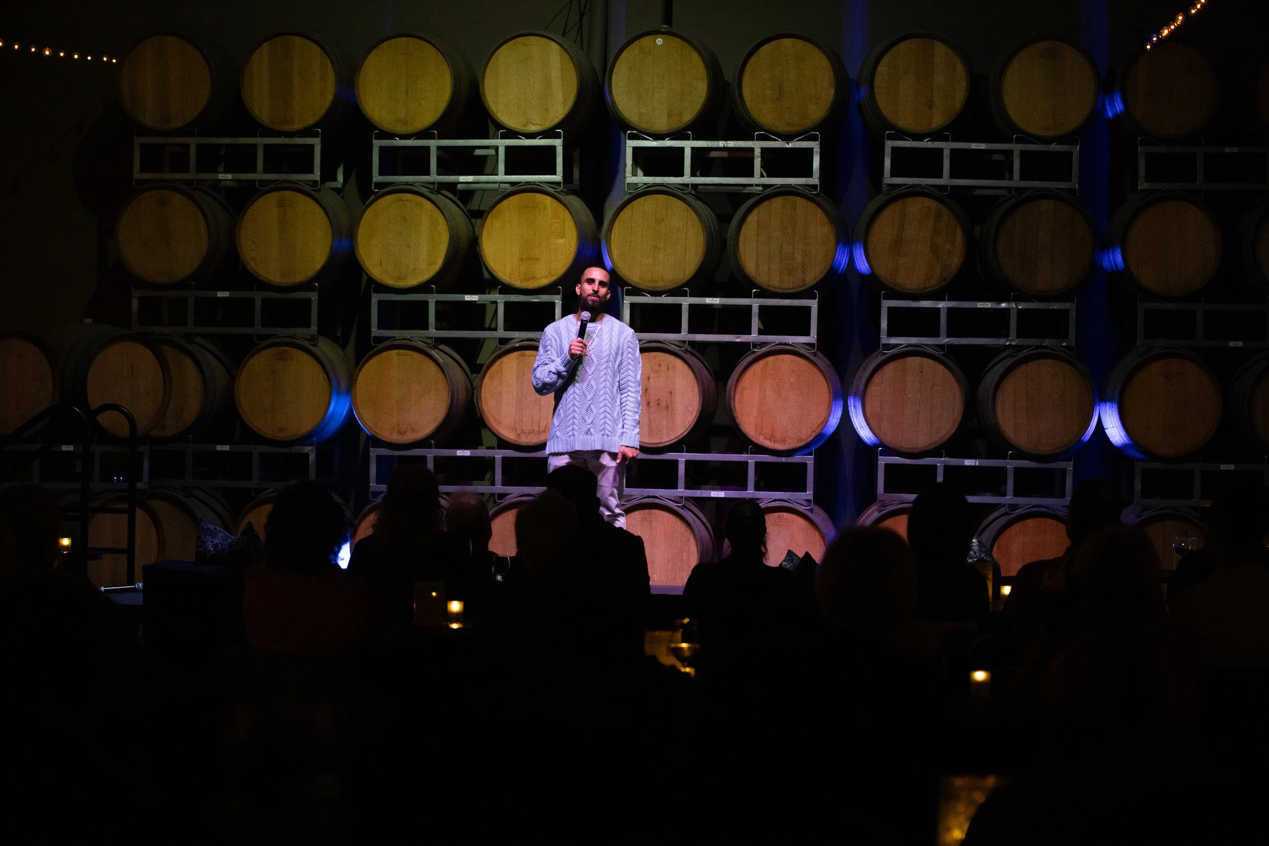 Person performing stand-up comedy in front of stacked wine barrels with audience seated in low lighting.