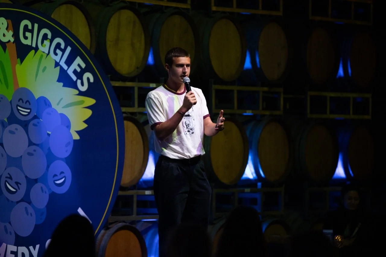 Person performing stand-up comedy with a microphone, in front of a backdrop with cartoon grapes and barrels stacked behind them.