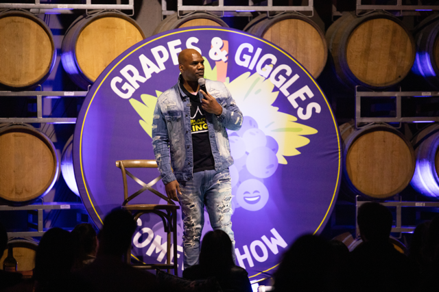 Comedian performing on stage at "Grapes & Giggles" comedy show, with wine barrels in the background.
