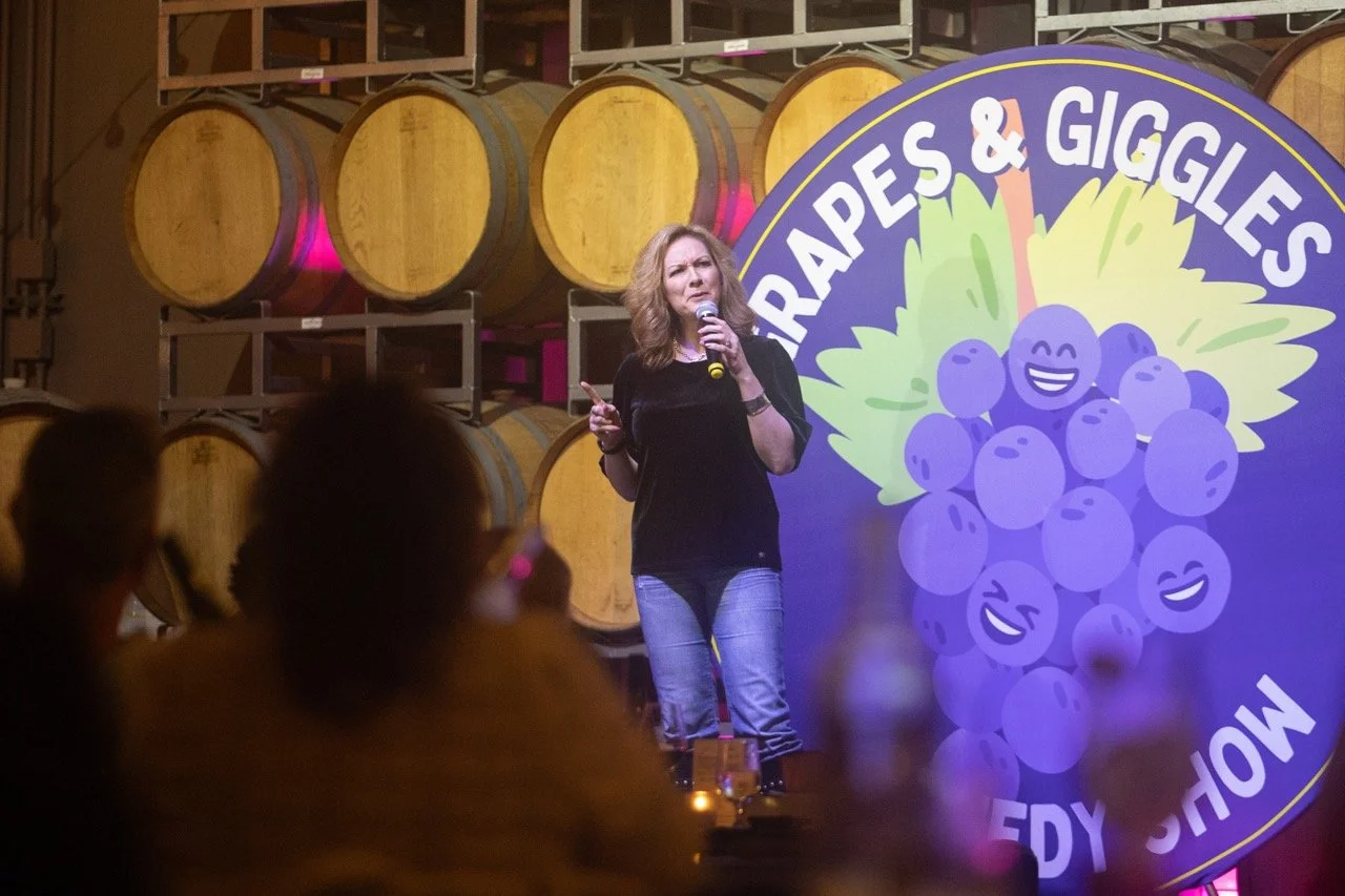 A woman performing stand-up comedy at an event called "Grapes & Giggles Comedy Show" with wine barrels in the background.