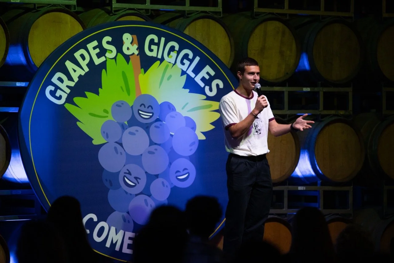 Comedian holding a microphone on stage at "Grapes & Giggles" comedy event with barrels in the background.