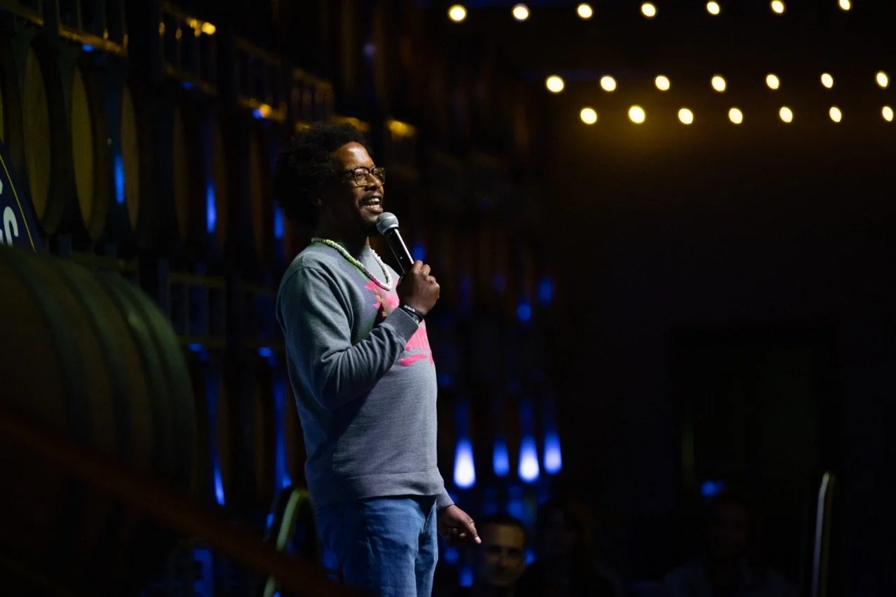 A man performing stand-up comedy on stage, holding a microphone, with dim lighting and string lights above. Barrels are visible in the background.