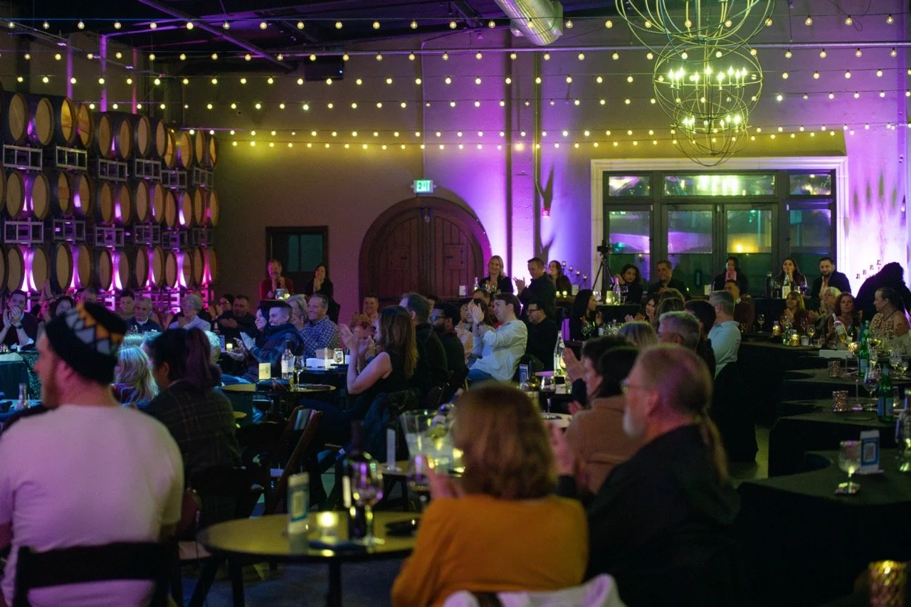 An indoor event with an audience seated at tables, surrounded by decorative lights and wine barrels on the walls.