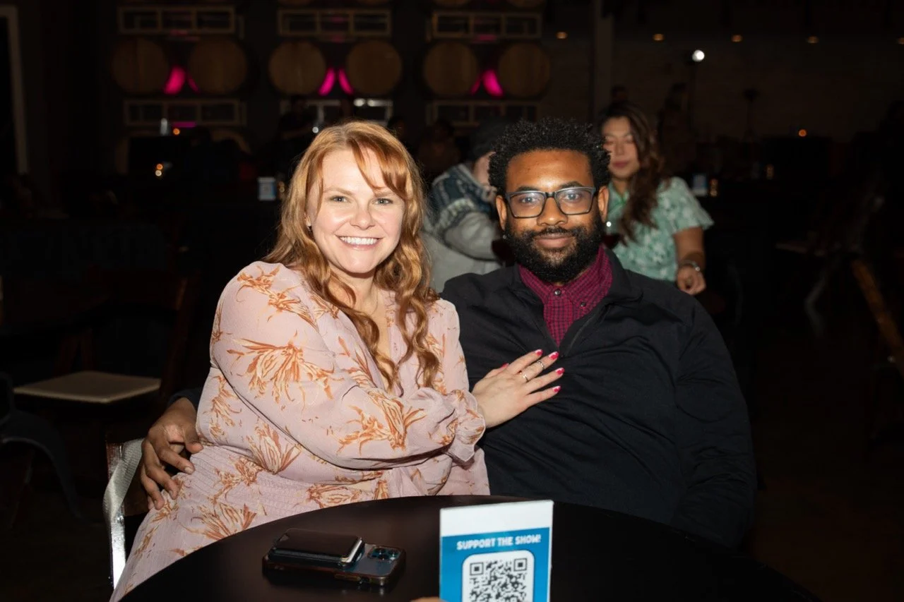A smiling man and woman sitting at a table in a dimly lit venue with barrels in the background. The woman has her hand on the man's shoulder. A mobile phone and QR code display are on the table.