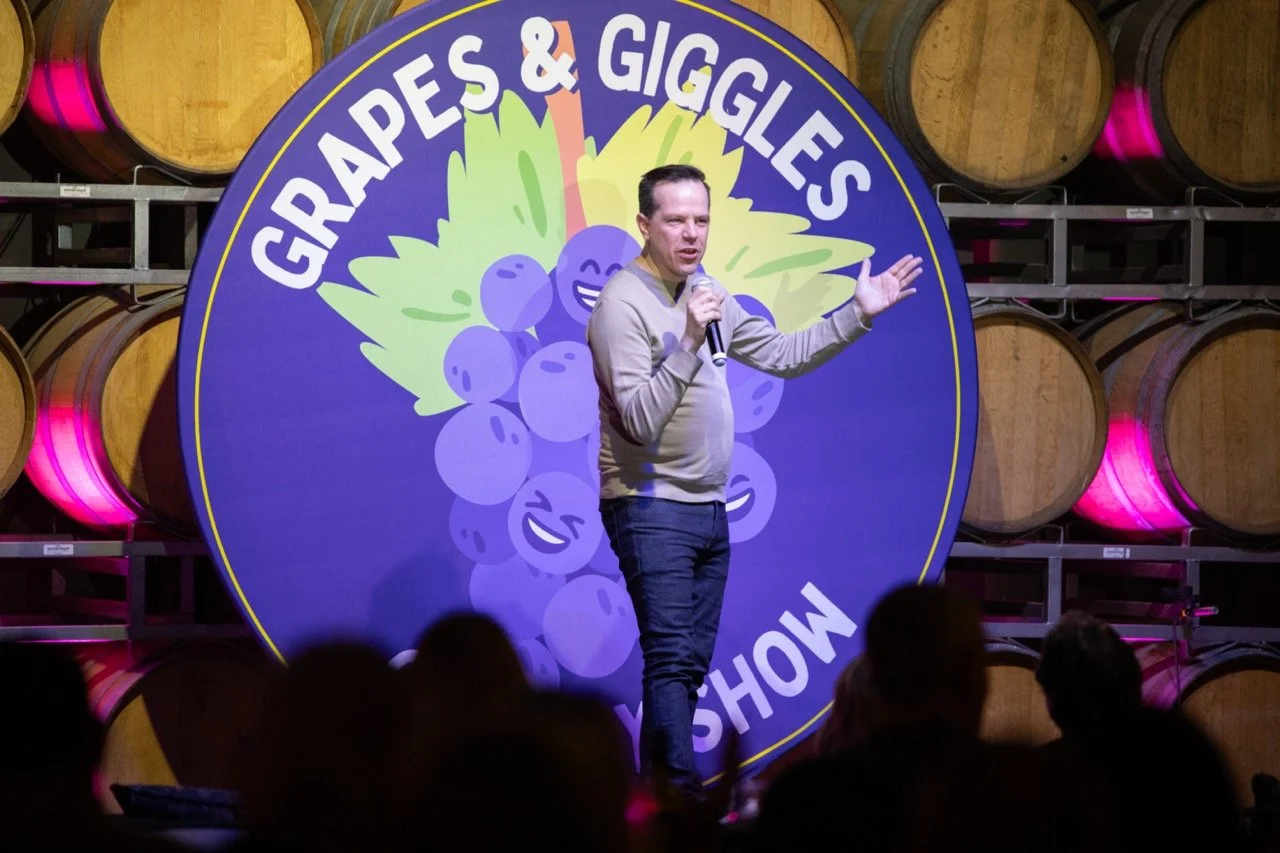 Comedian performing at Grapes & Giggles comedy show, wooden barrels in background.